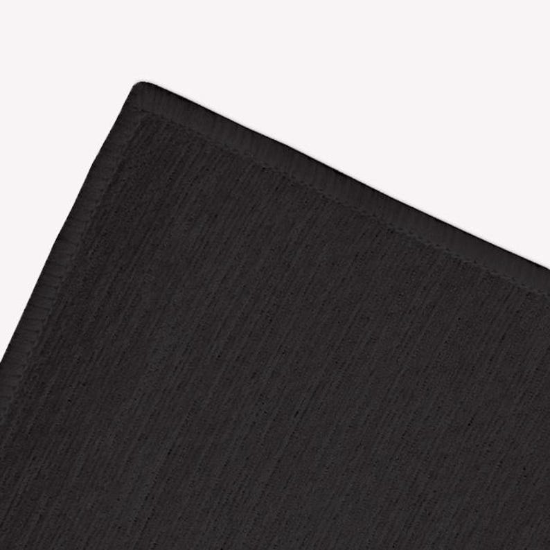 May include: A corner of a black rectangular rug with a textured surface. The rug has a stitched border and a solid black color. The rug is made of a soft material.