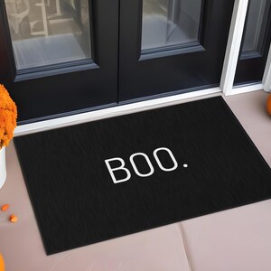 May include: Black rectangular doormat with the word "BOO." in white. The mat is placed in front of a black door, with pumpkins and orange flowers on either side, creating a festive Halloween scene.