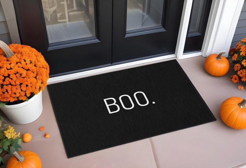 May include: Black rectangular doormat with the word "BOO." in white. The mat is placed in front of a black door, with pumpkins and orange flowers on either side, creating a festive Halloween scene.