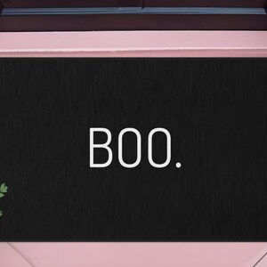 May include: Black rectangular doormat with the word "BOO." in white. The mat is placed in front of a door, with a pumpkin and greenery on either side, suggesting a Halloween theme.