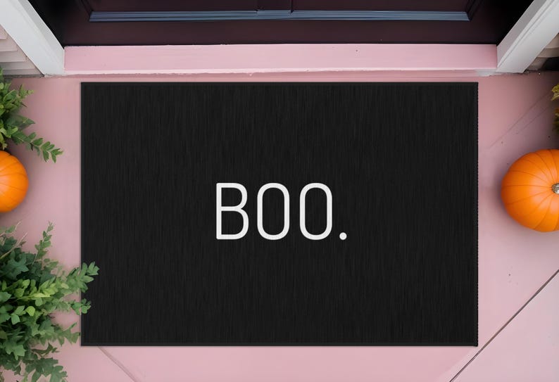 May include: Black rectangular doormat with the word "BOO." in white. The mat is placed in front of a door, with a pumpkin and greenery on either side, suggesting a Halloween theme.