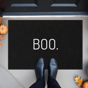 May include: A black doormat with the word "BOO" in white lettering. The mat is surrounded by orange pumpkins and fall leaves, creating a festive Halloween scene. Black shoes are visible.