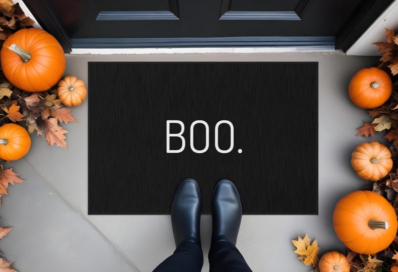 May include: A black doormat with the word "BOO" in white lettering. The mat is surrounded by orange pumpkins and fall leaves, creating a festive Halloween scene. Black shoes are visible.
