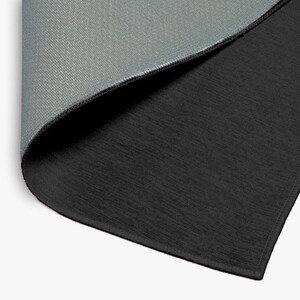 May include: A close-up view of a black and gray rug with a textured surface. The rug is folded over, revealing the gray underside. The edges are neatly finished with a dark border. The rug appears to be made of a durable material.