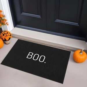 May include: Black doormat with the word "BOO." in white, surrounded by orange pumpkins and potted orange flowers, creating a festive Halloween scene.