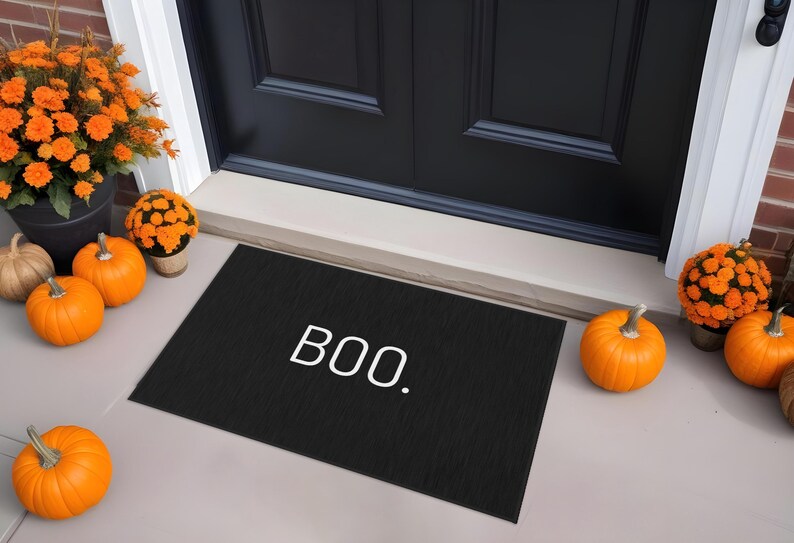 May include: Black doormat with the word "BOO." in white, surrounded by orange pumpkins and potted orange flowers, creating a festive Halloween scene.