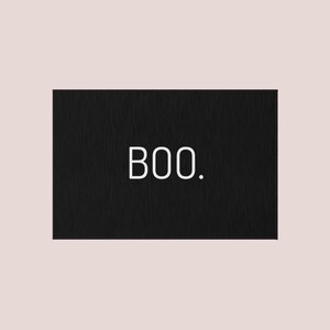 May include: A rectangular black sign with the word "BOO." in white, sans-serif font. The sign has a textured appearance and is set against a pale pink background. The design is simple and minimalist.