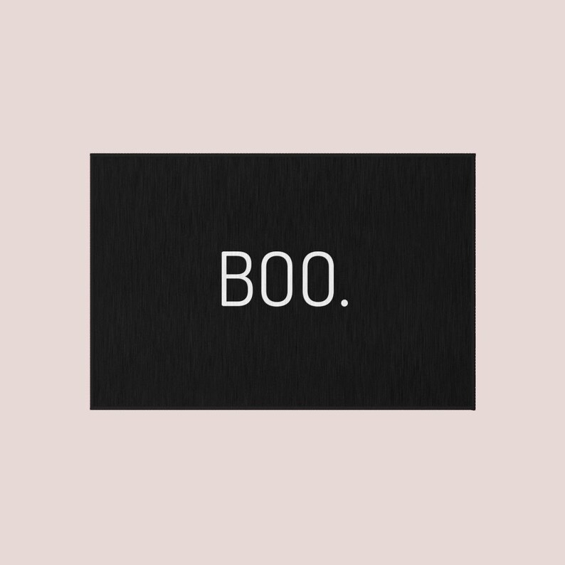 May include: A rectangular black sign with the word "BOO." in white, sans-serif font. The sign has a textured appearance and is set against a pale pink background. The design is simple and minimalist.
