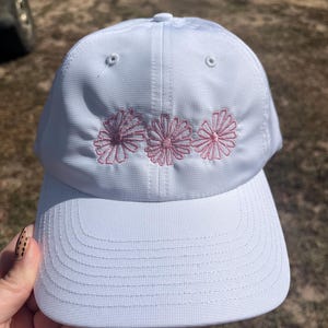 May include: A white baseball cap with three embroidered pink flowers across the front. The cap has a curved brim and a structured crown. The hat is held by a hand with a patterned nail design.