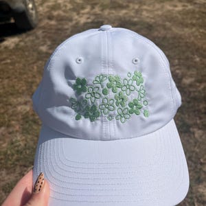 May include: A white baseball cap with a curved brim. The cap features an embroidered design of green flowers on the front. The cap has a button on top and eyelets for ventilation. The cap is made of a textured fabric.