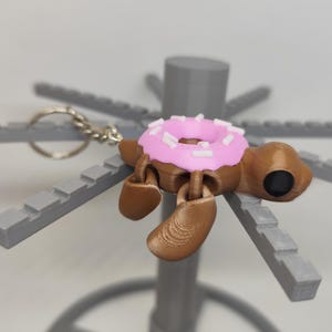 Op de afbeelding: Een bruine sleutelhanger in de vorm van een schildpad met een roze donut-schaal en witte hagelslag. De schildpad heeft een zilveren sleutelring. De poten van de schildpad zijn bruin en hij heeft een zwart oog.