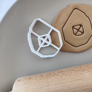 以下が含まれることがあります： 白いプラスチック製のクッキー型で、ダイヤモンドの形をしています。内側に小さなダイヤモンドがあります。クッキー型は白い表面に置かれており、その隣には、ダイヤモンドの形が切り抜かれた茶色の粘土があります。