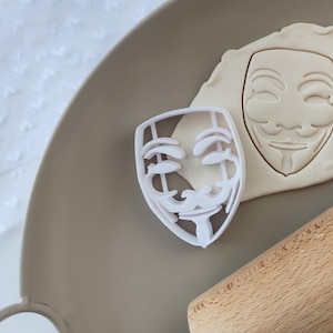 May include: White plastic cookie cutter shaped like the Anonymous mask. The mask has a white outline and a black interior. The cookie cutter is on a brown surface with a rolling pin.