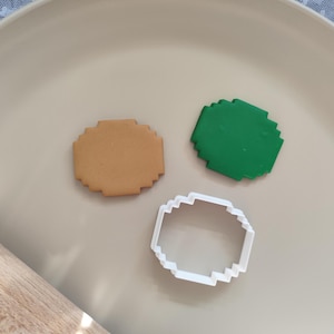 May include: A tan biscuit, a green piece of fondant, and a white biscuit cutter in a pixelated design are arranged on a beige plate. The biscuit cutter is in the shape of the biscuit and fondant. A rolling pin is visible in the lower left corner.