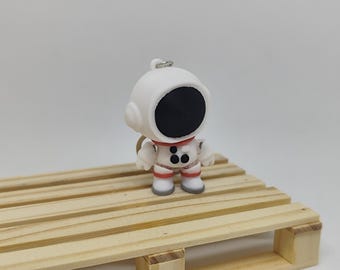 May include: A white astronaut figurine with a black visor and red accents. The astronaut is standing on a small wooden pallet. The figurine has a keychain attachment.