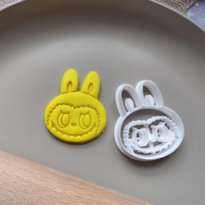 Peut inclure: Un emporte-pièce en forme de lapin jaune et un biscuit jaune. Le biscuit a un visage de dessin animé avec de grands yeux et un sourire. L'emporte-pièce est blanc et a le même design de visage de lapin.