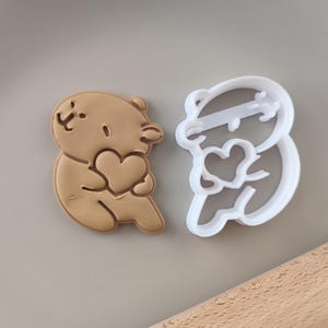 Puede incluir: Un cortador de galletas marrón en forma de oso sosteniendo un corazón. Un cortador de galletas de plástico blanco en forma de oso sosteniendo un corazón.