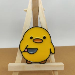 May include: Yellow cartoon chick holding a knife enamel pin. The pin is on a small wooden easel.