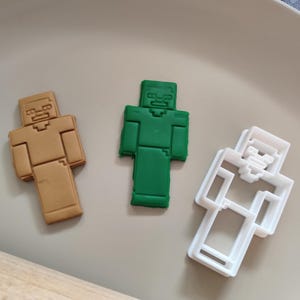 Può includere: Tre elementi a tema Minecraft: un biscotto color marrone, un biscotto color verde e un tagliabiscotti in plastica bianca. I biscotti e il tagliabiscotti hanno la forma di un personaggio di Minecraft. Gli oggetti sono su un piatto color marrone chiaro.