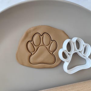 May include: A white plastic cookie cutter in the shape of a paw print. A brown clay paw print is next to the cutter.