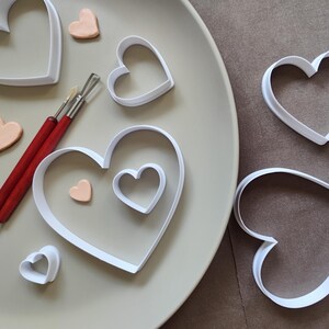 May include: A collection of white heart-shaped cookie cutters in various sizes, accompanied by two sculpting tools with red handles and several small, light peach-colored heart shapes made of clay, arranged on a smooth, light-colored surface.