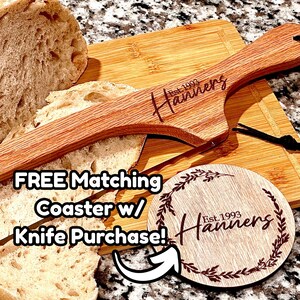 Könnte beinhalten: Ein Brotmesser aus Holz und ein passendes Untersetzer-Set. Das Messer ist mit "Hanners Est. 1993" graviert. Der Untersetzer ist rund mit einem floralen Design und demselben Text. Das Messer liegt auf einem Holzschneidebrett mit geschnittenem Brot.