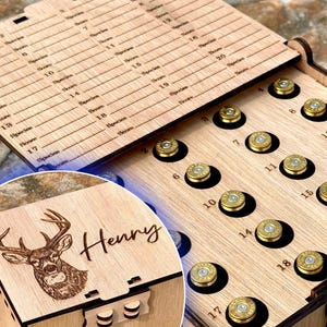 May include: A wooden box with a hinged lid, featuring a deer head engraving and the name "Henry." The box is designed to hold brass bullet casings in numbered slots. The lid has engraved lines and numbers for species and score.