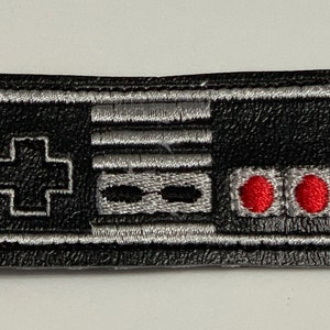 May include: Black leather keychain with a silver embroidered image of a classic video game controller. The controller has a directional pad, two buttons, and a start button.