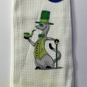 May include: A white waffle-weave kitchen towel featuring an embroidered gray meerkat wearing a green top hat, vest, and bow tie. The meerkat holds a green pipe and a walking stick. A blue circular tag reads The finest goods in the galaxy! IMPERIAL.