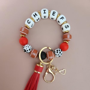 May include: A beaded keychain bracelet with the word "CHIEFS" spelled out in white letter beads. The bracelet includes football-shaped beads, red beads, and a red tassel. A gold-colored clasp and heart charm complete the design.