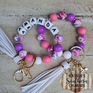 May include: Two beaded wristlet keychains featuring round silicone and floral patterned beads in shades of pink, purple, and white. One keychain includes white letter beads spelling AMANDA. Both have white leather tassels and gold hardware.