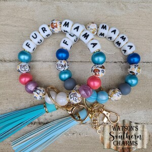 May include: Two beaded keychains featuring white letter beads spelling FUR MAMA, interspersed with multicolored round beads in blue, pink, purple, and grey, along with patterned beads showing dog illustrations and light blue leather-like tassels.