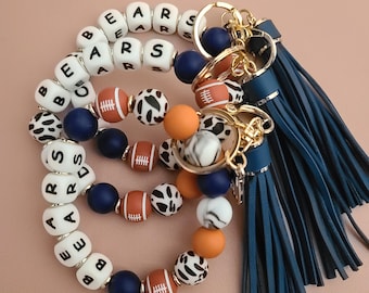 May include: Three beaded keychains spelling "BEARS" with white letter beads. The keychains feature navy, orange, football-shaped, and cow-print beads. Each keychain has a navy tassel and gold-coloured hardware.