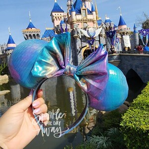 May include: A pair of iridescent blue and purple Minnie Mouse ears with a large bow. The ears are in front of a blue and white castle with a blue sky in the background. The text "Sparkle the Day Away" is written on the headband.