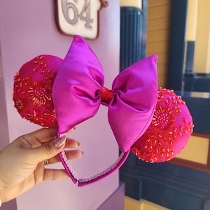 May include: A pair of magenta mouse ears with a large satin bow. The ears are embellished with red beading and embroidery. The headband is white and the bow is a vibrant magenta color.