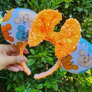 May include: Orange and blue Mickey Mouse ears headband with a pumpkin and Mickey Mouse design. The headband has a large orange bow made of sequins.
