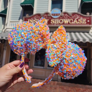 May include: Orange Minnie Mouse ears with iridescent sequins. The ears are in the shape of a bow and are held in a hand. The background is a building with the text "Disney Showcase".