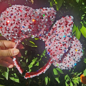 May include: Pink sequined headband with large ears, adorned with red, white, and iridescent sequins. The headband is held against a backdrop of green foliage and oranges, creating a festive and eye-catching accessory.