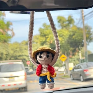 Puede incluir: Un adorno de coche de ganchillo hecho a mano que representa a un personaje con sombrero de paja, chaleco rojo, pantalones cortos azules y una faja amarilla. El adorno cuelga de un espejo retrovisor, con un fondo borroso de una escena callejera.