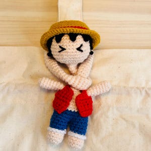 Puede incluir: Muñeco de ganchillo de un personaje con sombrero amarillo con banda roja, pelo negro, chaleco rojo y pantalón azul. El muñeco está sujeto a una bolsa de mano color crema. Los brazos del muñeco están cruzados sobre el pecho.