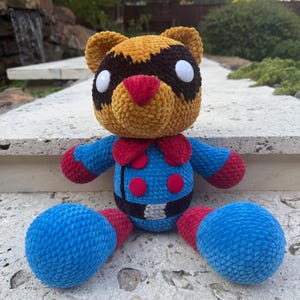 以下が含まれることがあります： 手作りのぬいぐるみクマ。 クマは、白い目、赤い鼻、赤いアクセントが付いた青い体を持つ茶色とタン色の頭をしています。 クマの足は青く、黒いベルトが付いています。魅力的でカラフルなぬいぐるみです。