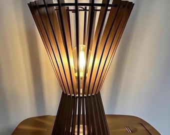 Può includere: Una lampada da tavolo in legno dal design unico. La lampada presenta un paralume a doghe aperte marrone scuro che si allarga da una base più stretta. La lampada è illuminata, con la lampadina visibile attraverso le doghe. La lampada è su un tavolo di legno.