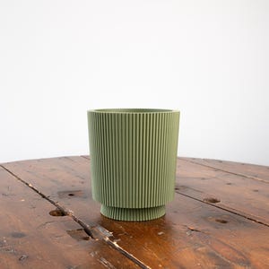 Peut inclure: Un pot en céramique vert à nervures avec une base large. Le pot est posé sur une table en bois.