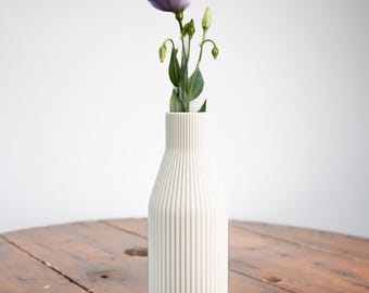 May include: A single purple flower with green leaves in a white ceramic vase with vertical lines. The vase is sitting on a wooden table.
