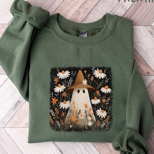 May include: Military green sweatshirt featuring a whimsical ghost wearing a brown hat, surrounded by white daisies and orange flowers. The ghost design is centered on the front of the sweatshirt. The text "MILITARY GREEN" is at the top.