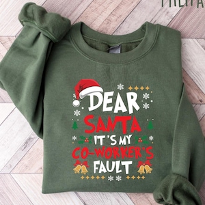 May include: Military green sweatshirt with the text "DEAR SANTA IT'S MY CO-WORKER'S FAULT" in white and red. The design includes a Santa hat, snowflakes, and bells, perfect for the holiday season.