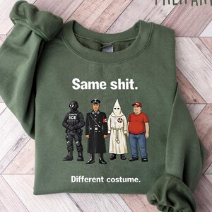 May include: Military green sweatshirt with the text "Same shit. Different costume." and illustrations of four figures in different uniforms. The sweatshirt is folded on a light wood-grain surface.