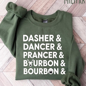 May include: A military green sweatshirt with white text that reads "DASHER & DANCER & PRANCER & BOURBON & BOURBON &". The sweatshirt has a crew neck and long sleeves. The words are stacked vertically. The background is a light wood pattern.