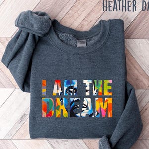 May include: Heather dark gray sweatshirt with the text "I AM THE DREAM" in colorful block letters. The sweatshirt is folded, showing the sleeves and neckline. The text features a portrait with vibrant colors.