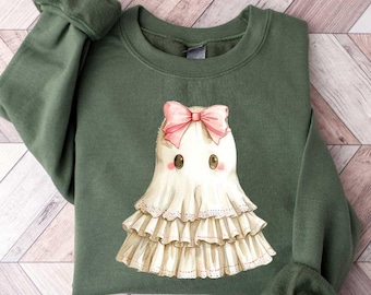 May include: An olive green sweatshirt featuring a watercolor illustration of a friendly ghost. The ghost has a pink bow, rosy cheeks, and a ruffled dress. The sweatshirt has long sleeves with ribbed cuffs.